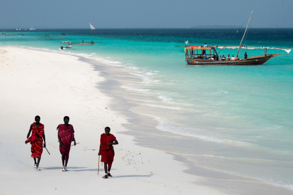 Trois Massaïs marchant sur une plage de Zanzibar avec un dhow tanzanien en arrière-plan, scène typique d’un voyage ou d’un voyage organisé en Tanzanie.