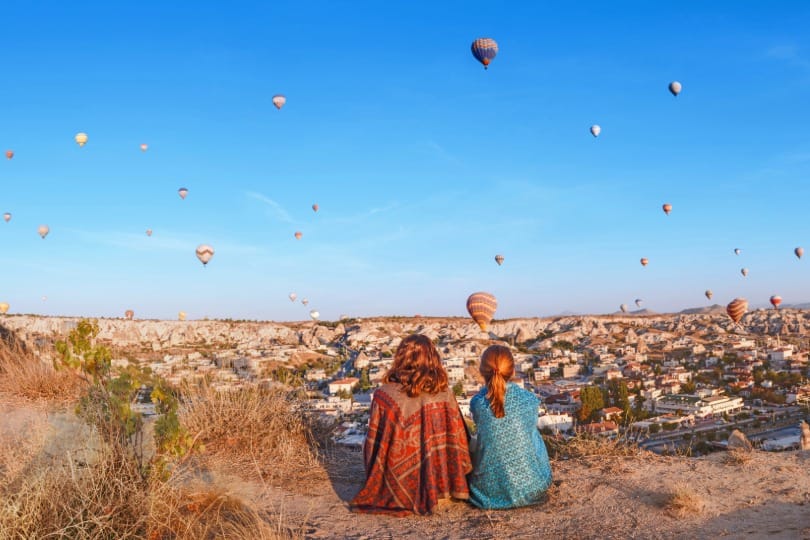 Deux voyageuses assises devant la vallée de Cappadoce en Turquie, regardant des montgolfières s'élever, parfait pour un voyage organisé ou un voyage romantique.